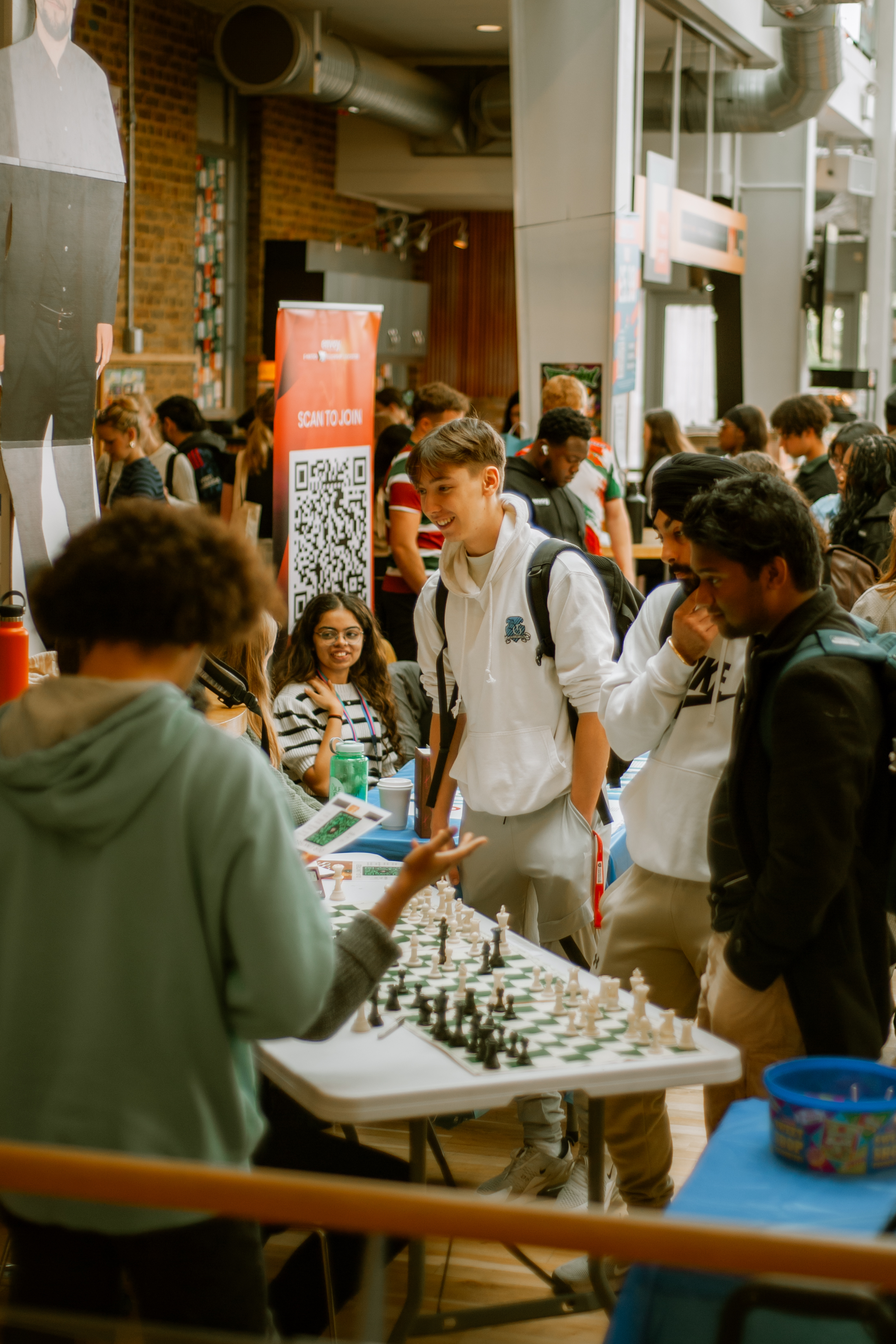 Photo from societies Fair, of a group of student stood around a table with chess